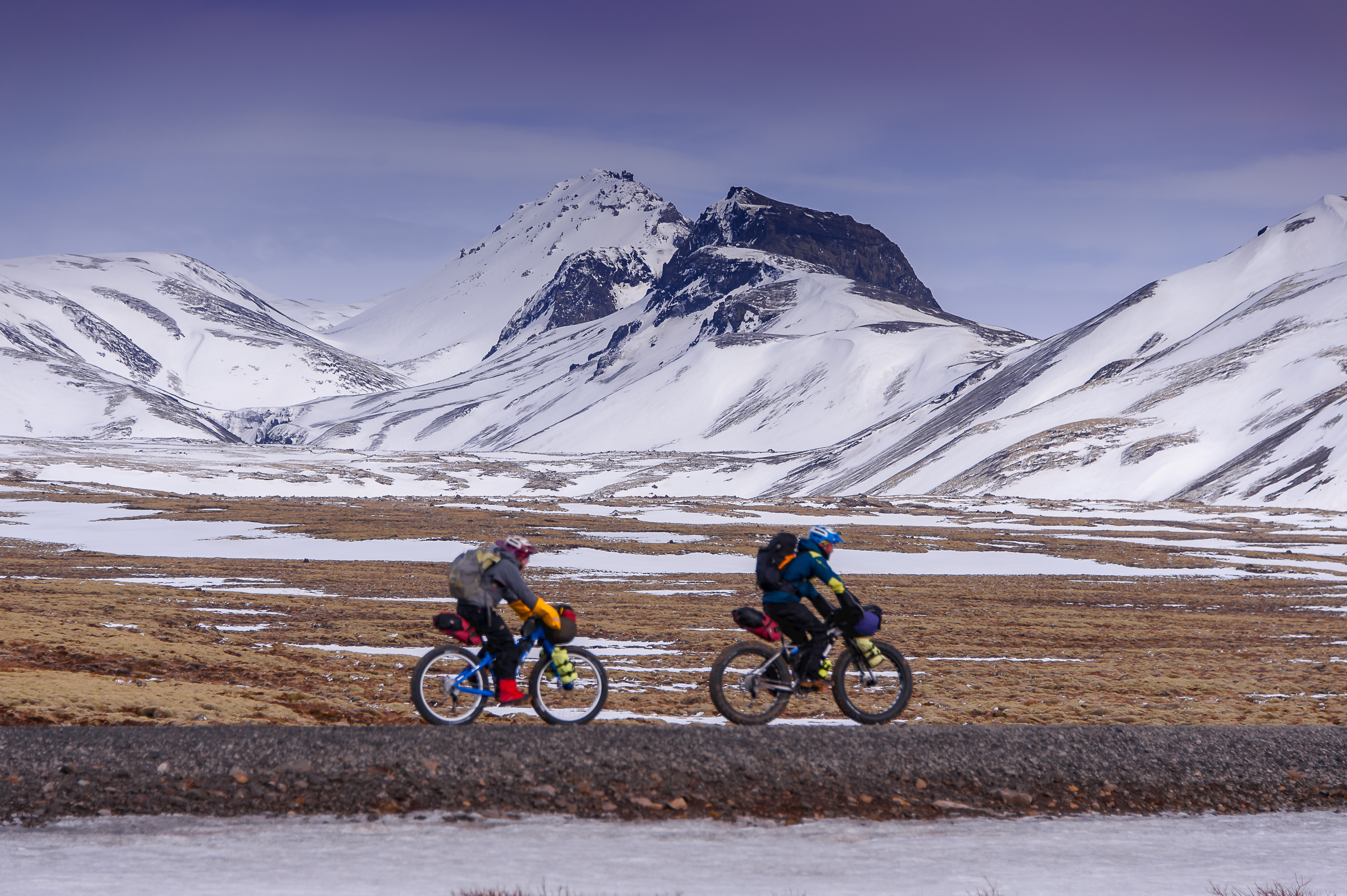 Two people cycle through a snowy, mountainous landscape in Iceland